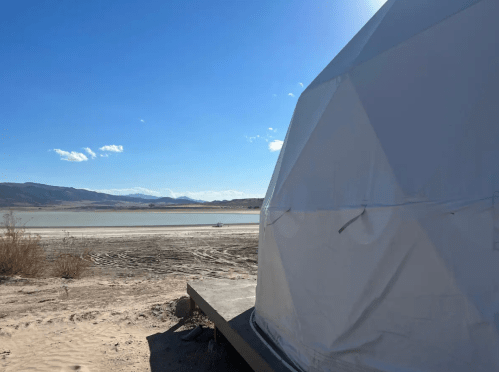 A white geodesic dome on a sandy shore, with mountains and a clear blue sky in the background.